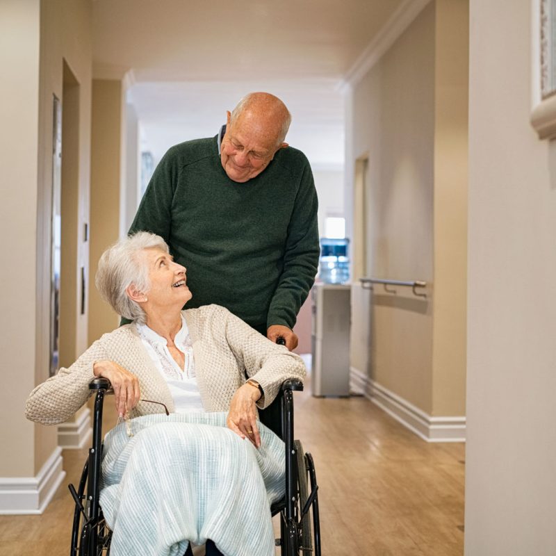 An older man pushing an elderly woman in a wheelchair and they're smiling at each other.